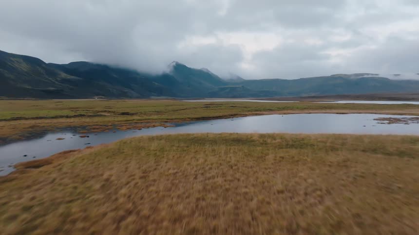An aerial moving shot descends over a large calm lake into a green and brown grassy plain with dark foggy mountains towering in the distance beneath a cloudy sky