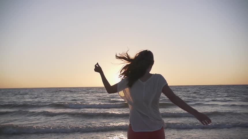 A woman running towards the water twirls around at sunset