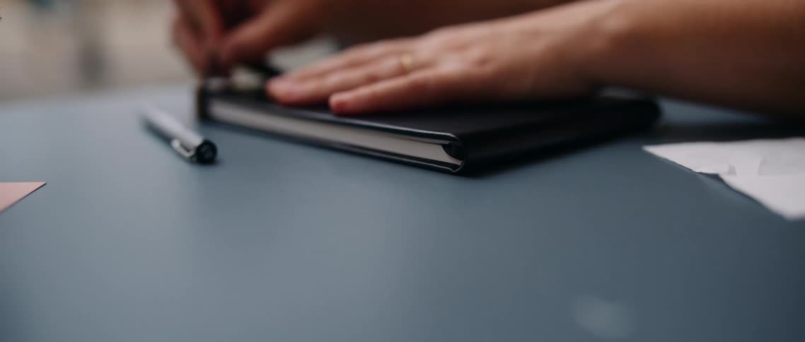 Closeup of a person closing a notebook on a desk with a pen nearby