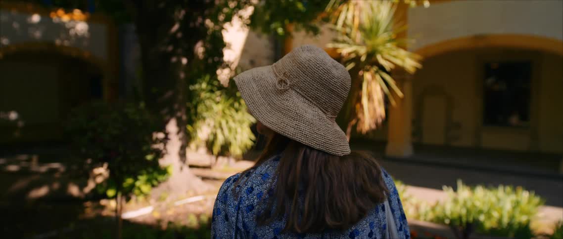 Woman in straw hat gazes at sunlit garden with mediterranean architecture in the background