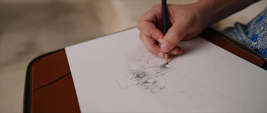 A close view shows a persons hand using a black pencil to draw a detailed technical sketch onto white paper placed on a brown leather surface