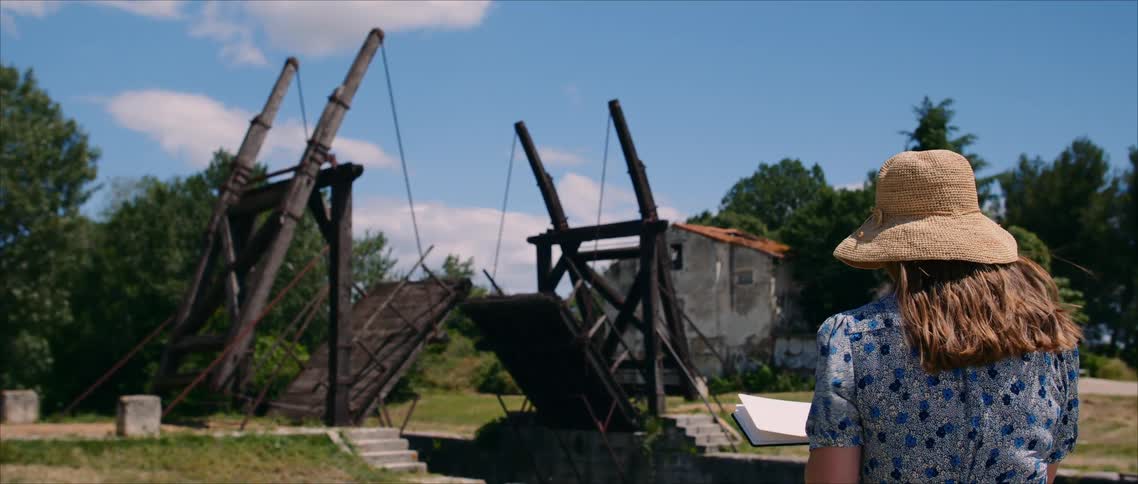 A rear view of a woman in a straw hat and blue patterned dress shows her holding a tablet or notebook while looking toward an old wooden drawbridge raised against a bright blue sky with a dilapidated stone building on the right
