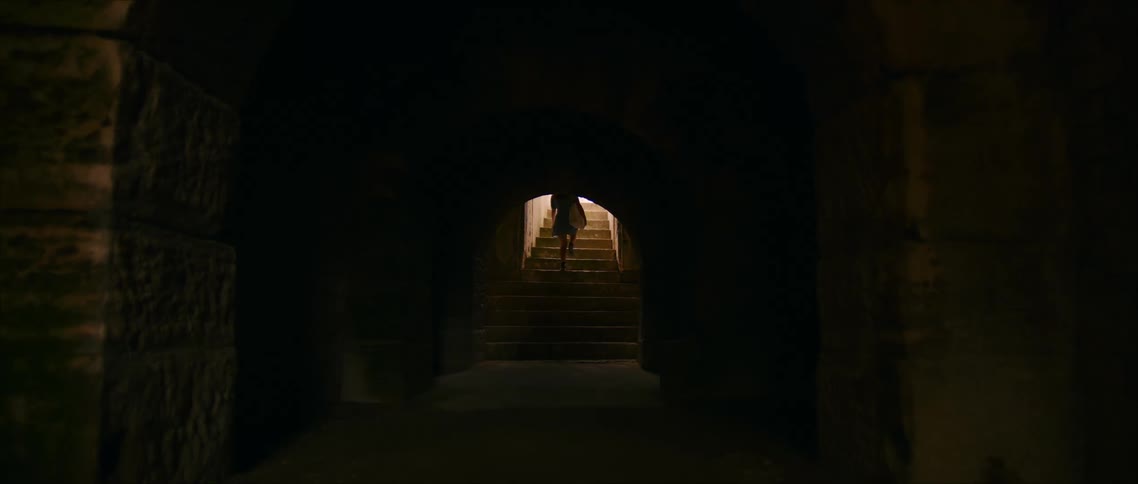 Figure ascends a lit staircase while leaving a dark stone corridor