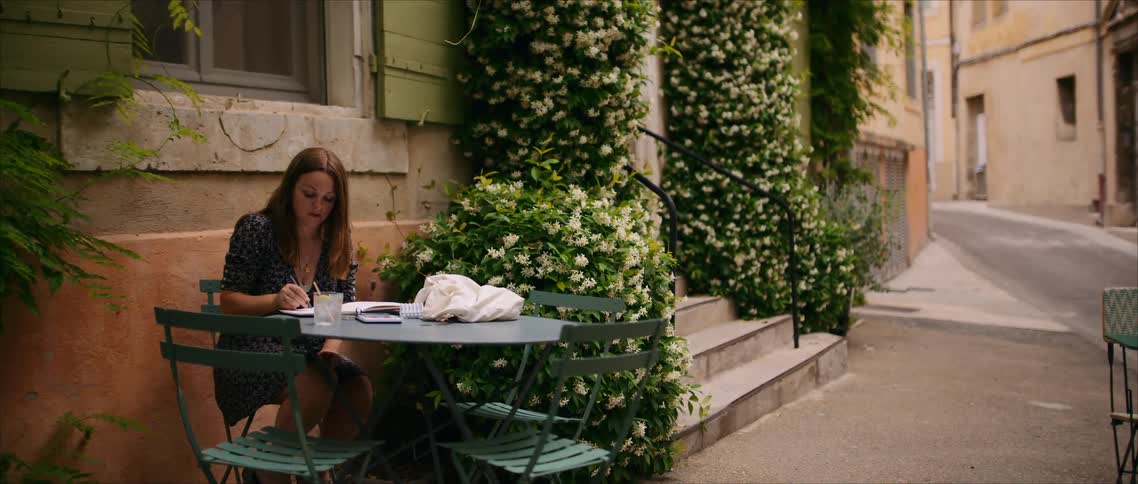 Woman sitting at an outdoor table writing in a notebook beside white flowers