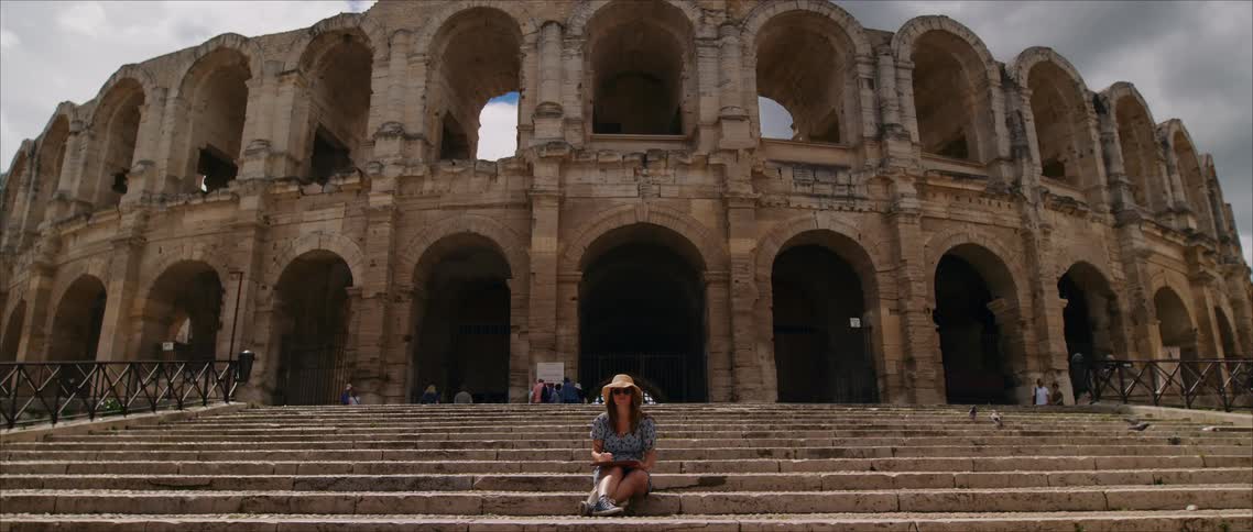 Man sits on steps facing roman amphitheater with cloudy sky in the background