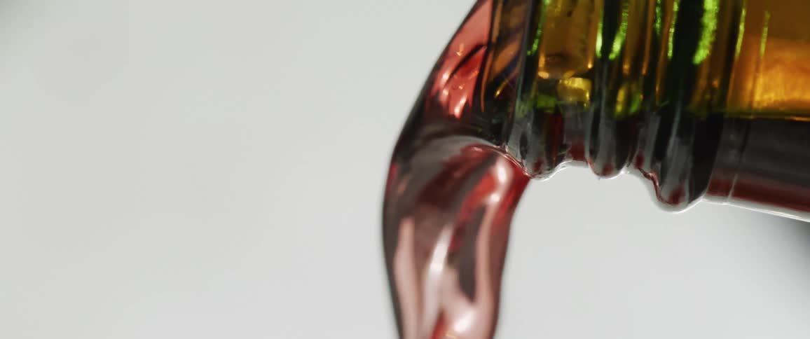 A closeup shot focuses on a thick dark reflective liquid with a green tint pouring in a slow stream from the serrated edge of a container against a bright white background