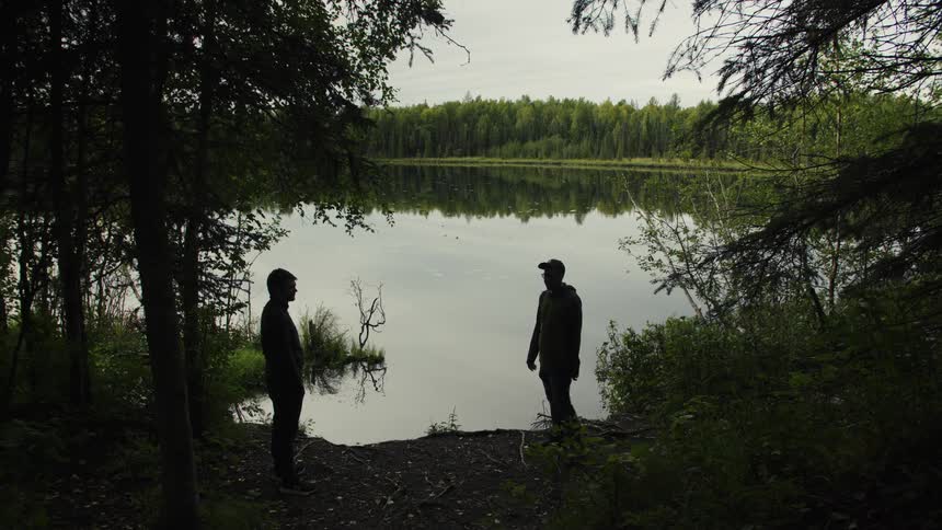 Figures observe calm lake surrounded by dense forest under cloudy sky