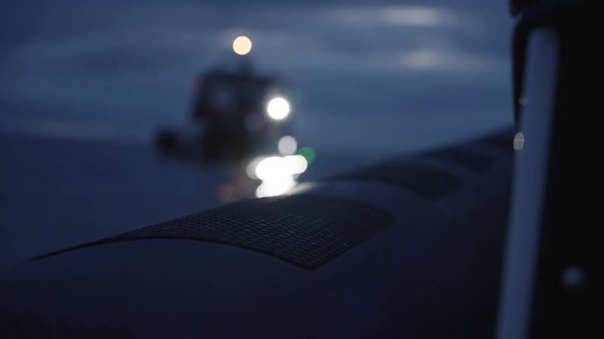 A lifeguard boat approaches another vessel with its lights on cutting through the dark waters under the night sky as reflections shimmer across the oceans surface