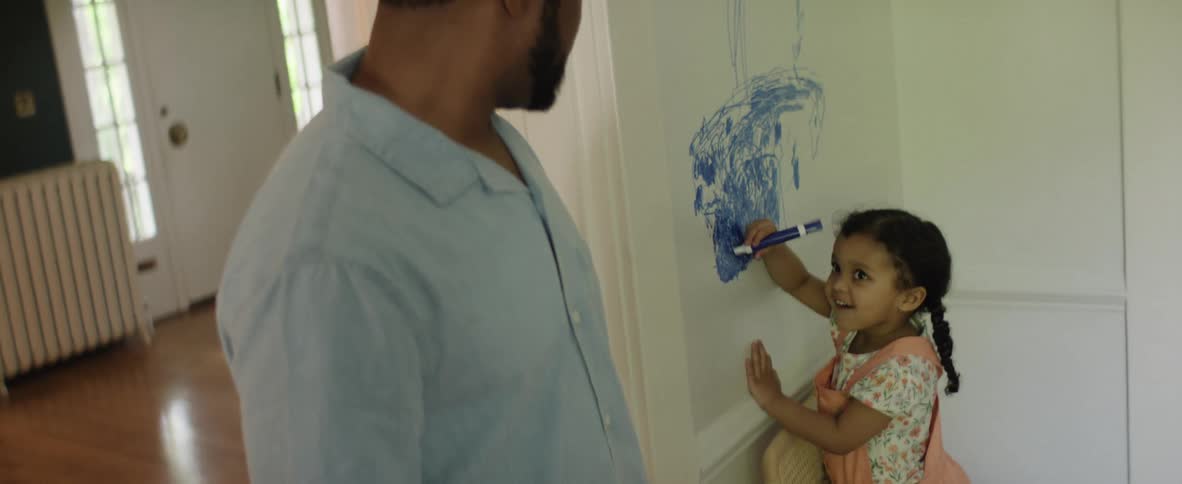 A little girl with a braid concentrates on coloring a big blue scribble on the wall with a marker as a man in a light shirt looks on