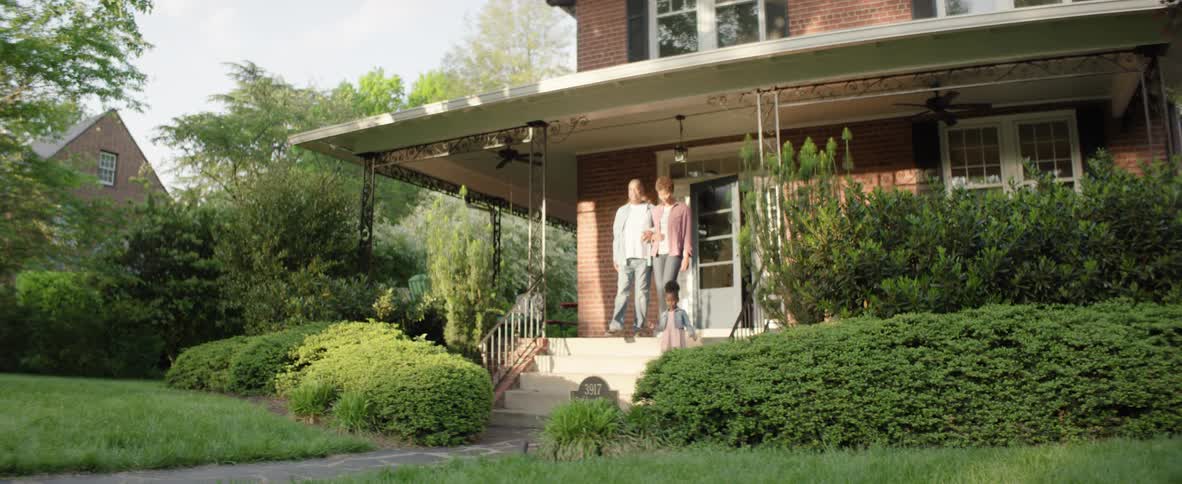 A man and a woman walk down the steps of a brick house porch as a young boy in an orange shirt and a young girl in a denim jacket and pink dress run past them across the green lawn