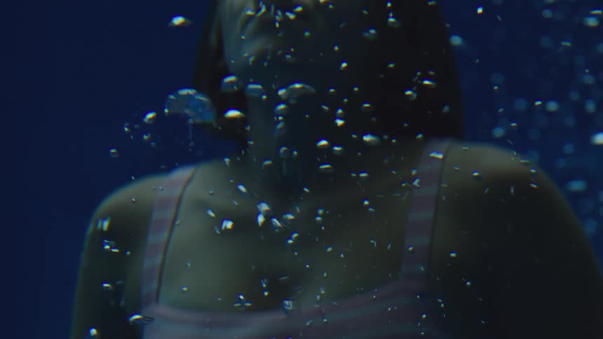 A woman in a pink tank top and white striped straps underwater with air bubbles rising past her against a dark blue background in a moody atmosphere