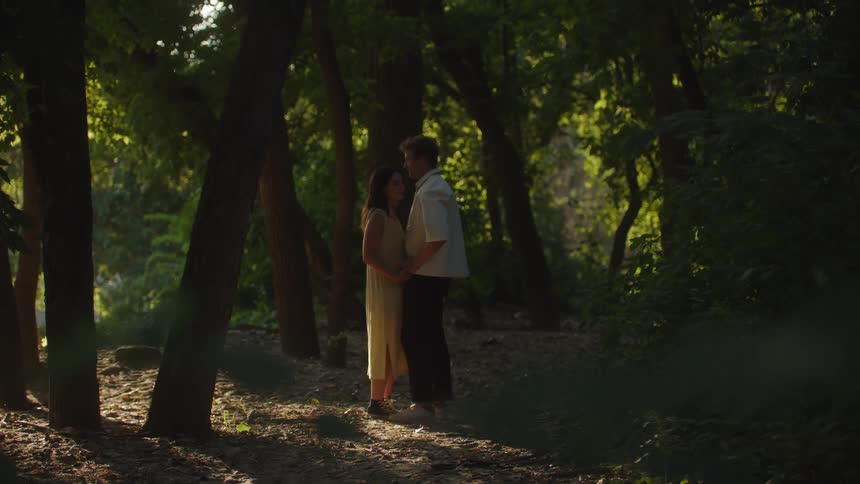 A female and male are standing close holding hands in a sunlit forest area