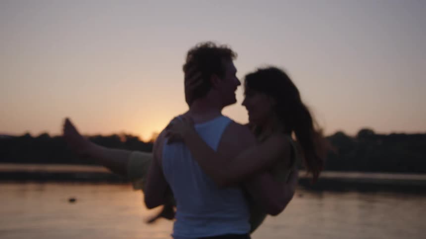 A man in a white tank top carries a woman in a pale dress in his arms as they embrace and kiss near the water with the sun setting behind them creating a dark silhouette