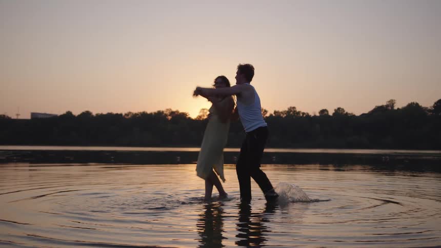 A couple dances and embraces in shallow water as the sun sets casting warm hues across the scene