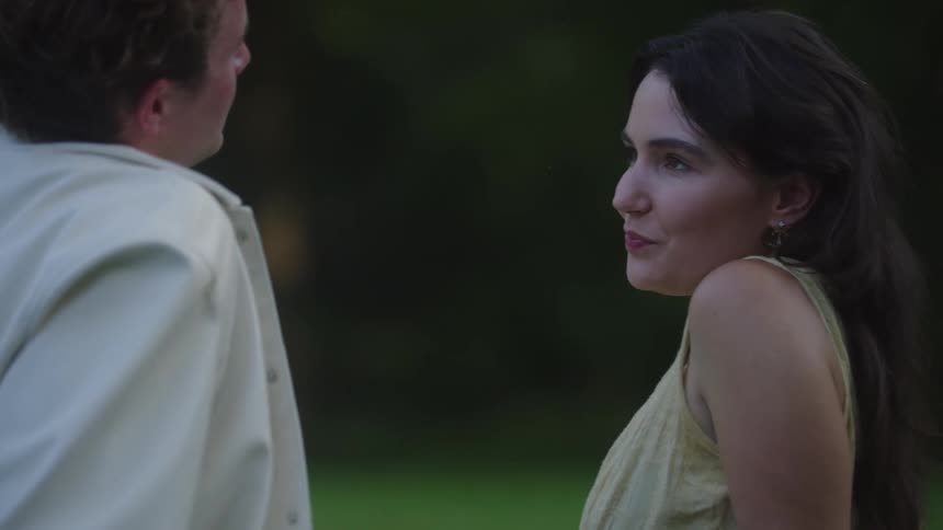 A young couple sits closely together on a sunlit grassland at midday sharing a romantic moment the young woman smiles warmly as she leans in gently touching her head to the mans and caressing his cheek surrounded by golden light and open fields