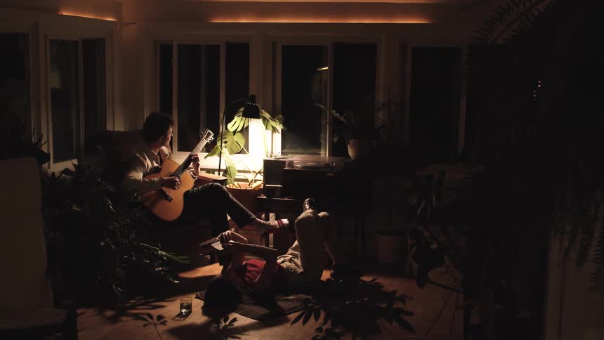 A man sitting in a chair plays an acoustic guitar for a woman relaxing on the floor next to him with a bright lantern illuminating the cozy plant filled sunroom at night