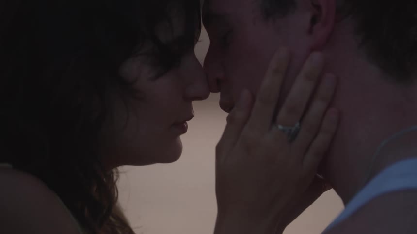 A young couple stands in shallow water sharing a romantic kiss as gentle waves lap around them and soft light reflects off the surface