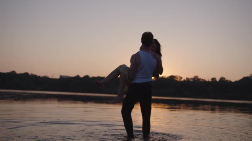 Man holds woman in his arms while standing in a lake at sunset