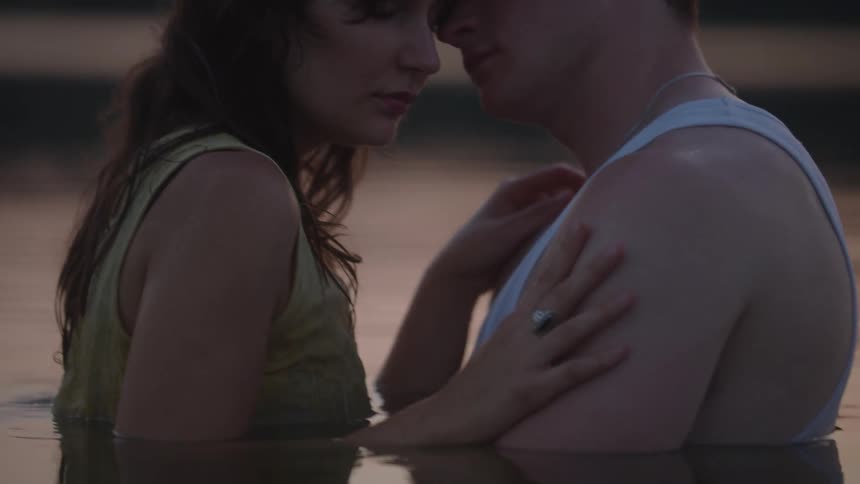 A couple stands forehead to forehead by the water their eyes soft and expressions tender