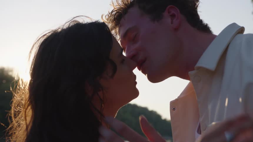 A closeup view shows a man and woman kissing passionately with the womans hand gently touching the mans face and the sun setting brightly behind them