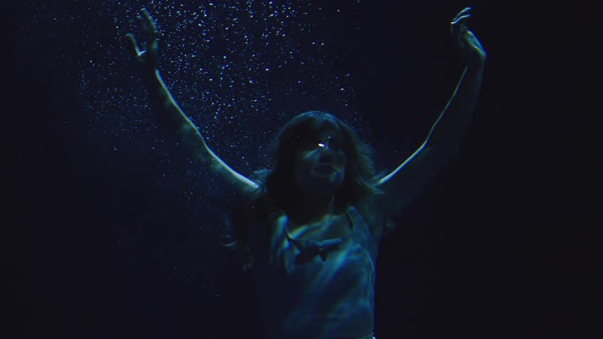 A woman in a pale tank top with her hair floating slowly descends underwater in a dark blue void with a stream of air bubbles rising above her head