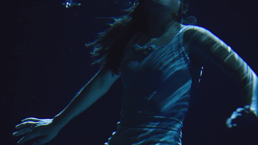 A woman in a light blue top with her hair flowing moves and turns underwater in a dark blue environment as light reflections ripple across her body