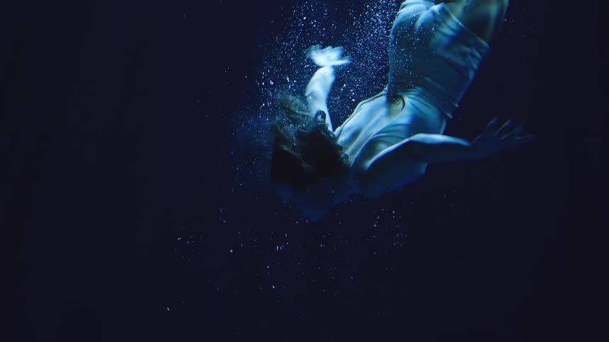 A woman in a pale blue swimsuit dives and turns underwater with her arms extended as a trail of bubbles follows her in a dark blue and moody environment