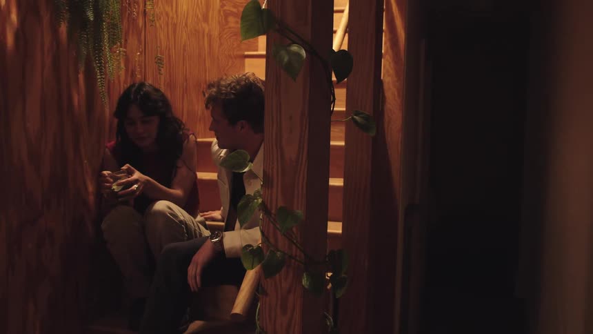 A woman sits on the lower steps of a wooden staircase holding a drinking glass to her head then a man walks over and sits beside her and they begin talking while facing each other