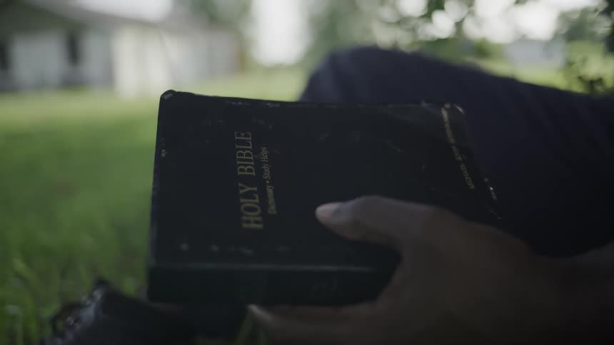 Hand holds a blackcovered bible as the person rests on the grass