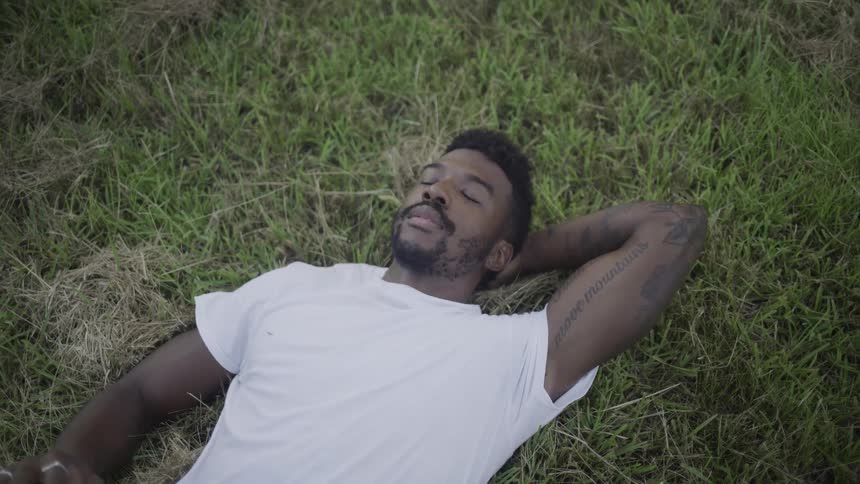 Black man wearing a white shirt lying down on the grass outdoors during day time