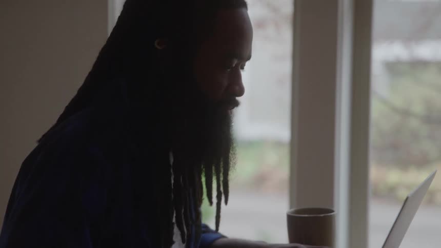 Man works on a laptop by a window with a notebook and mug on the table