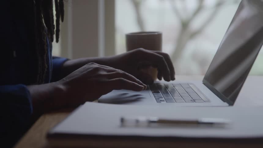 A closeup of a person with dreadlocks shows their hands quickly typing on a laptop keyboard that rests on a desk next to a cup with a window and natural light in the background