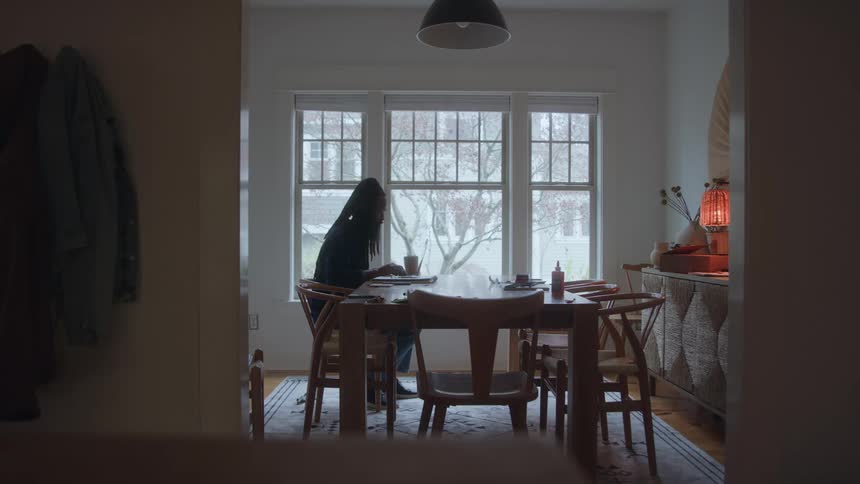 A woman with long dark hair leans over a large wooden table in a dining room while using a laptop as snow falls outside the wide window on a dark and moody day
