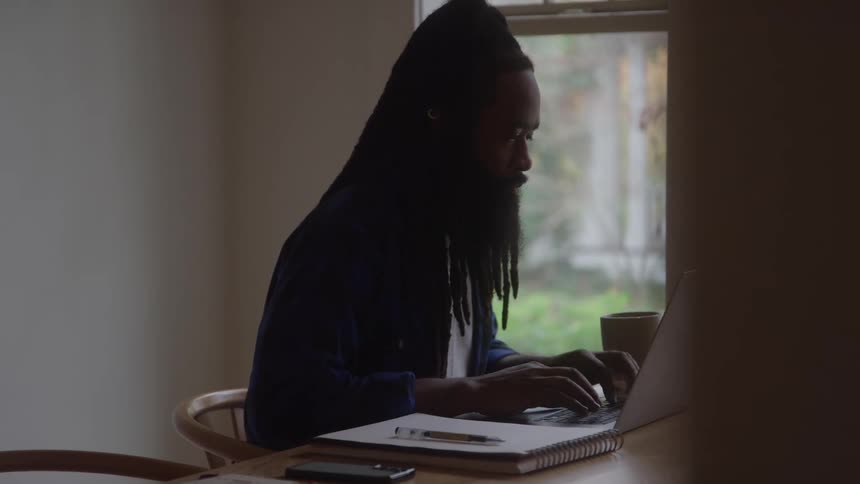 A man with long dark dreadlocks wearing a dark shirt sits at a wooden table using a silver laptop and drinking from a mug in a dimly lit room with a large window in the background