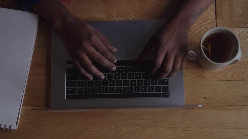 Person types on laptop over wooden surface with notebook on the left and tea mug on the right