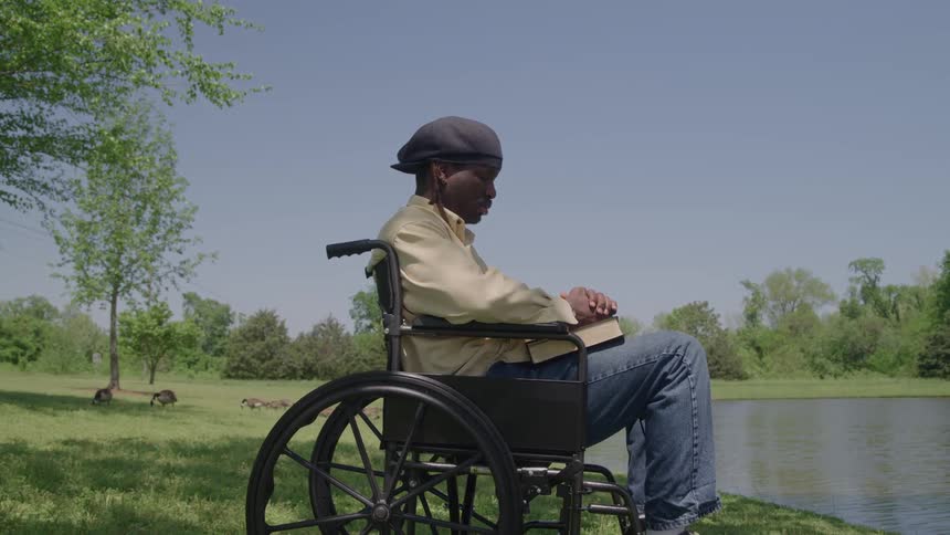 The video shows an older man in a wheelchair sitting by a small pond in a grassy parklike area he is holding a book and looking out over the water seemingly reading or in contemplation