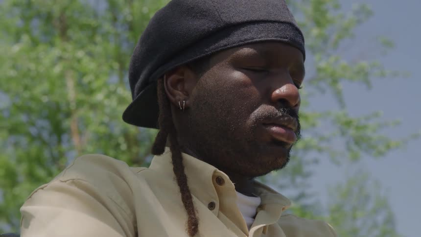 Face of a black man wearing a black hat and a white shirt with is eyes closed outdoors during day time