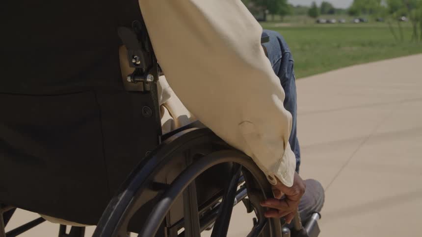 A black man in a wheelchair moves along a sunlit paved path his hand gripping the push rim with steady focus