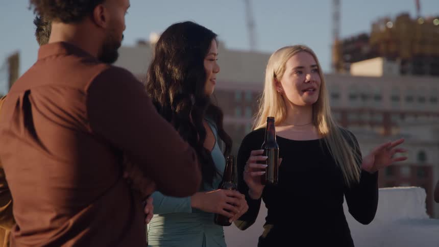 Three people chat while holding bottles on a rooftop at sunset