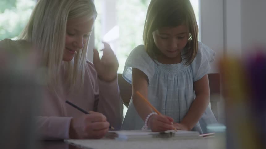 A woman with blonde hair wearing a pink top writes in a notebook next to a young girl in a light blue dress who is also writing with an orange pencil at a table near a bright window