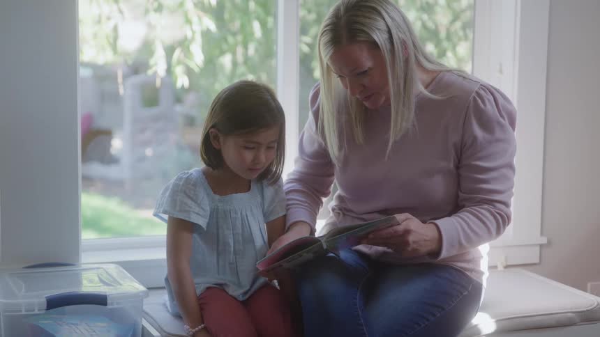 Mother reads a book beside her daughter sitting near a sunlit window