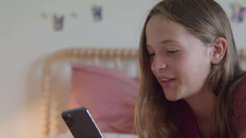 A close view shows a smiling young woman with long brown hair wearing a pink shirt looking down and typing on a black smartphone while lying in a bright bed