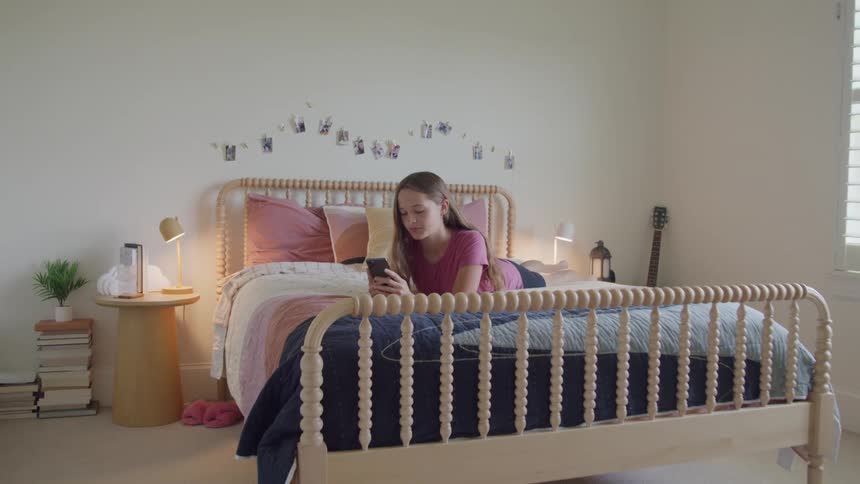 A teenager lies on a bed looking at a phone in a decorated bedroom