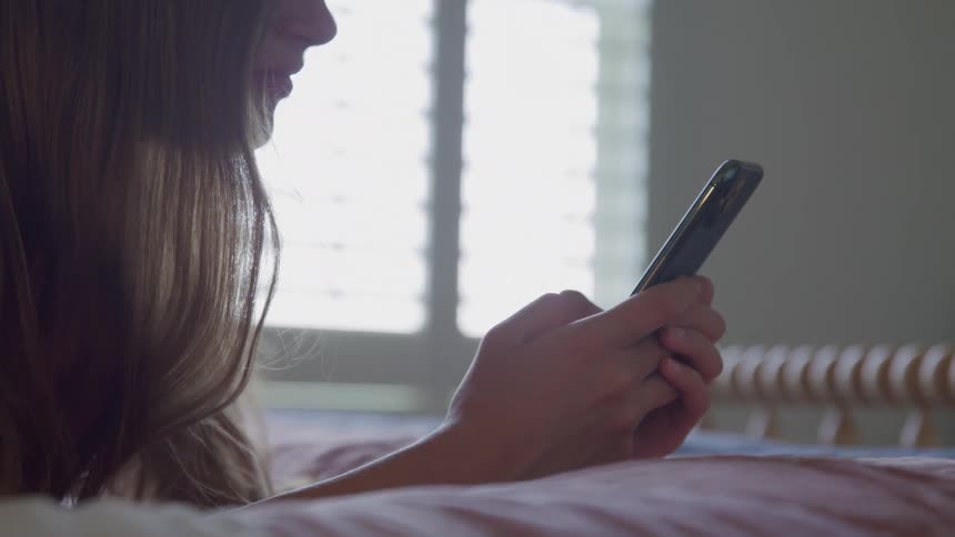 Girl lying on the bed holds a phone while looking at the screen
