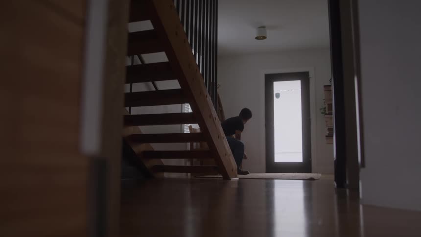 Young man walking down a staircase and then putting on shoes and a jacket before walking out of the door during day time
