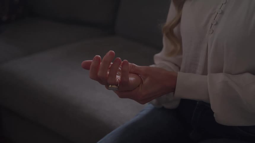 A woman sits on a sofa gently holding and rubbing her own wrist with her opposite hand