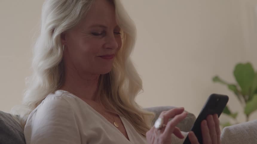 Blonde woman with wavy hair interacts with smartphone seated on couch with plant in the background