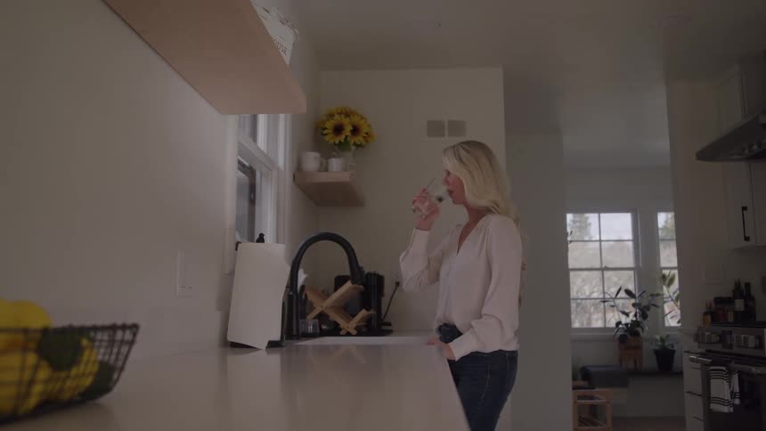 Person drinks water by the sink in welllit kitchen with wooden shelves and sunflower vase