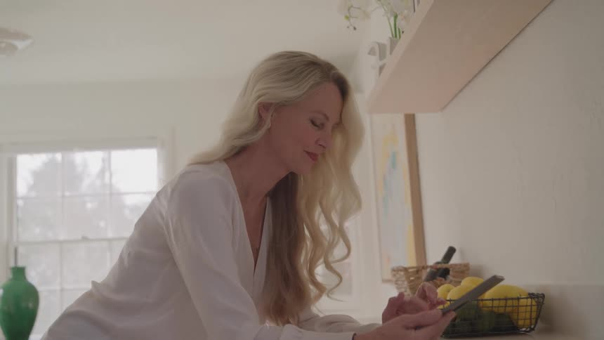 Blonde woman leans on white kitchen counter while interacting with smartphone next to fruit basket