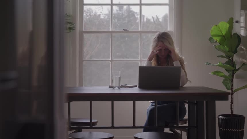 Woman holds head with hands while using laptop on wooden table in room lit by window